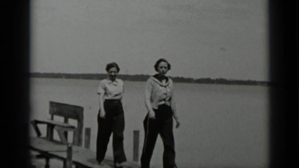 Two women walk toward the camera on a wooden pier at Clear Lake, California, with a bench, posts, still water, and shoreline in view.
