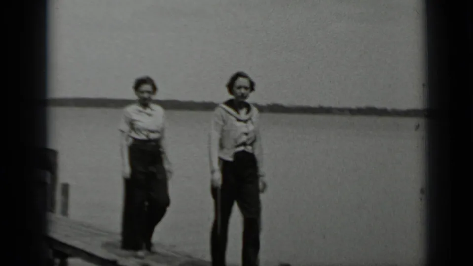 Two women in period clothing on a wooden pier at Clear Lake, California, with calm water and a low shoreline behind them.