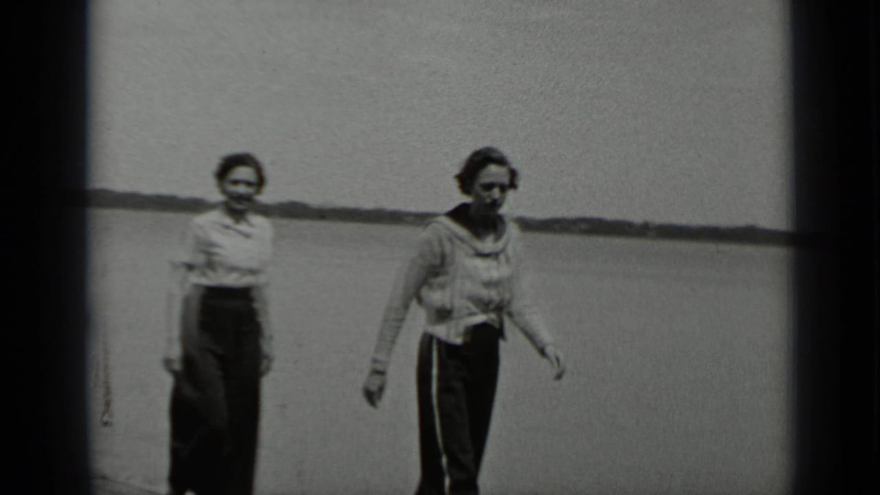 Two women walk closer to the camera on a pier beside open lake water.