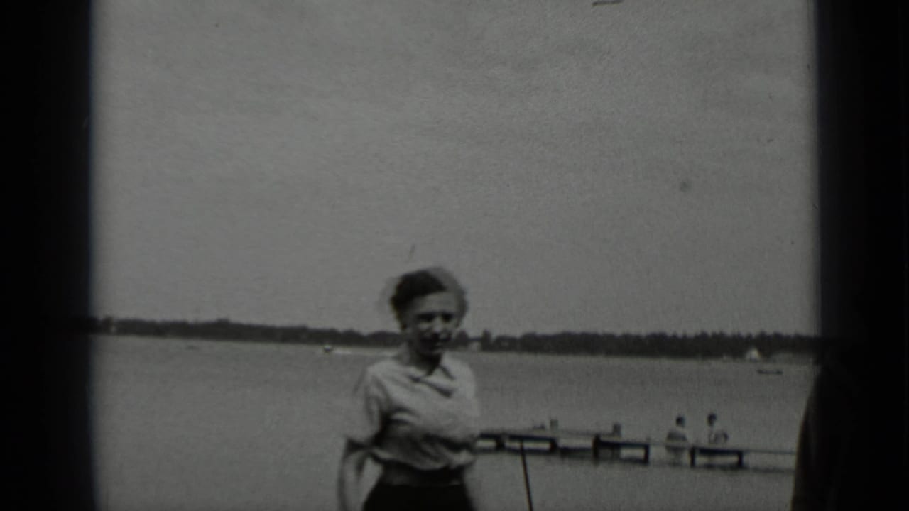 At Clear Lake in 1939, a woman in a light blouse walks near the camera with a wooden pier and calm lake behind her.