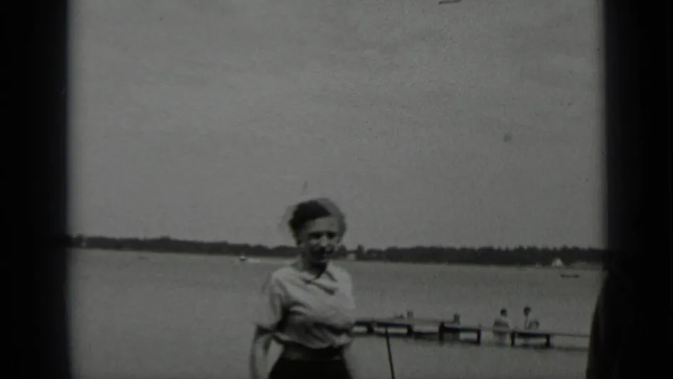 At Clear Lake in 1939, a woman in a light blouse walks near the camera with a wooden pier and calm lake behind her.