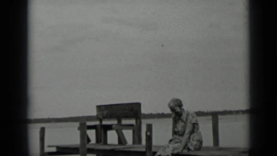 A woman sits on the dock at Clear Lake, with a wooden bench or pier structure and low shoreline behind her.