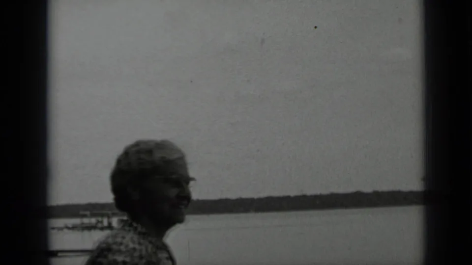 Older woman in patterned clothing smiles near Clear Lake, California, with calm water, dock structures, and a distant shore visible.
