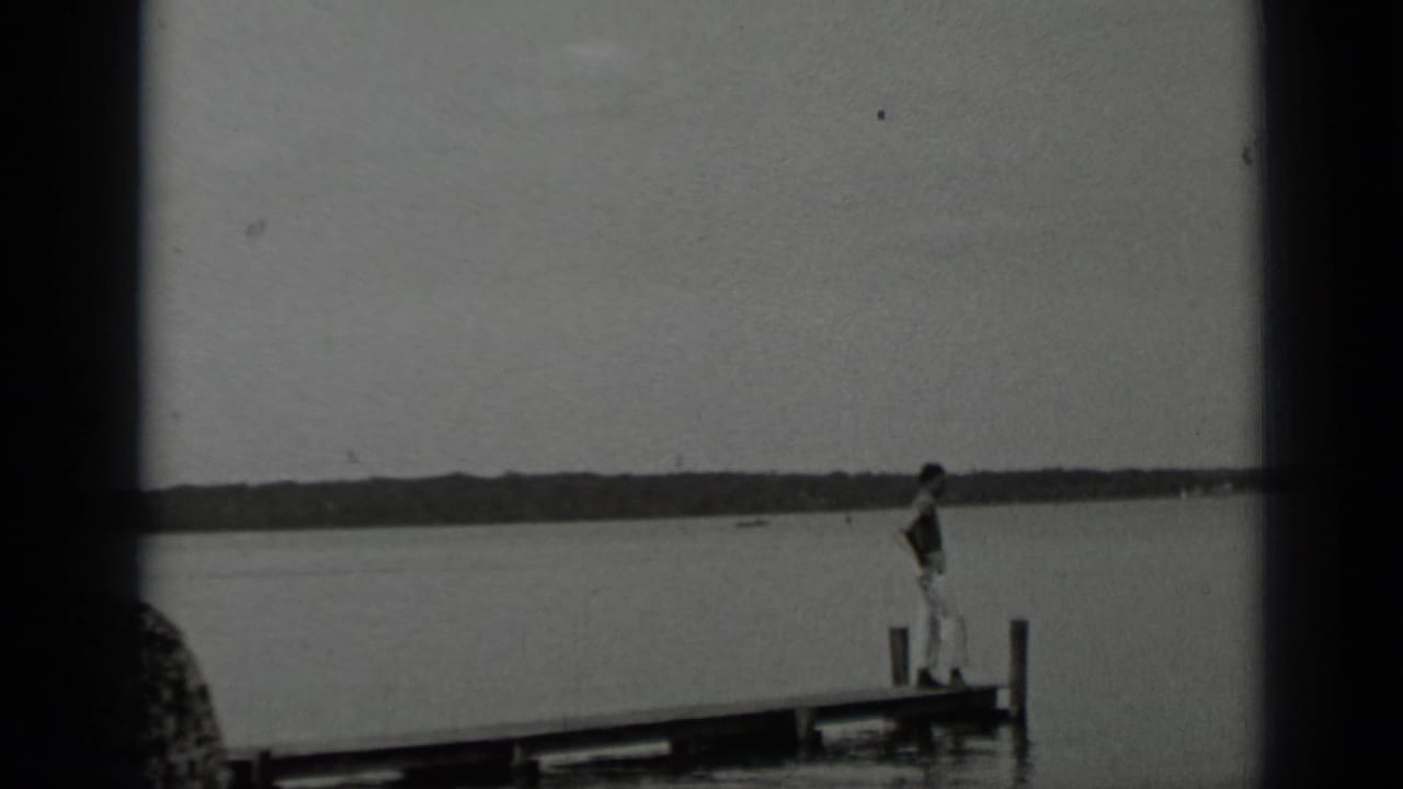Young man in light clothing walks along a wooden dock at Clear Lake, framed by calm water and a low distant treeline.