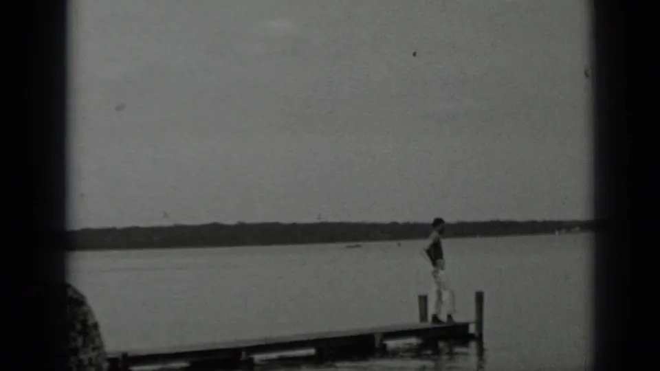 Young man in light clothing walks along a wooden dock at Clear Lake, framed by calm water and a low distant treeline.