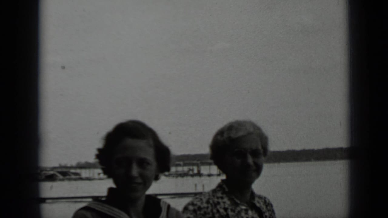 A younger woman and an older woman stand near the Clear Lake shore with dock structures, water, and distant shoreline behind them.