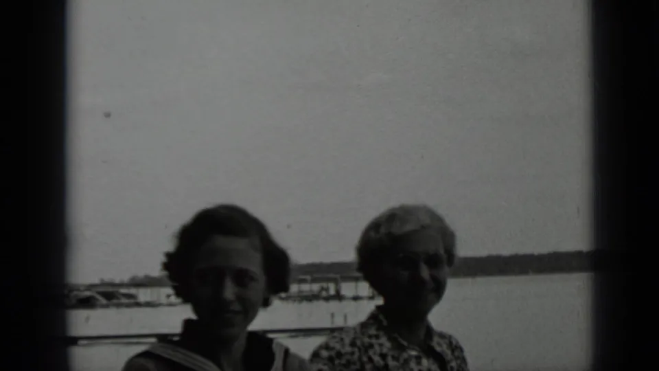 A younger woman and an older woman stand near the Clear Lake shore with dock structures, water, and distant shoreline behind them.