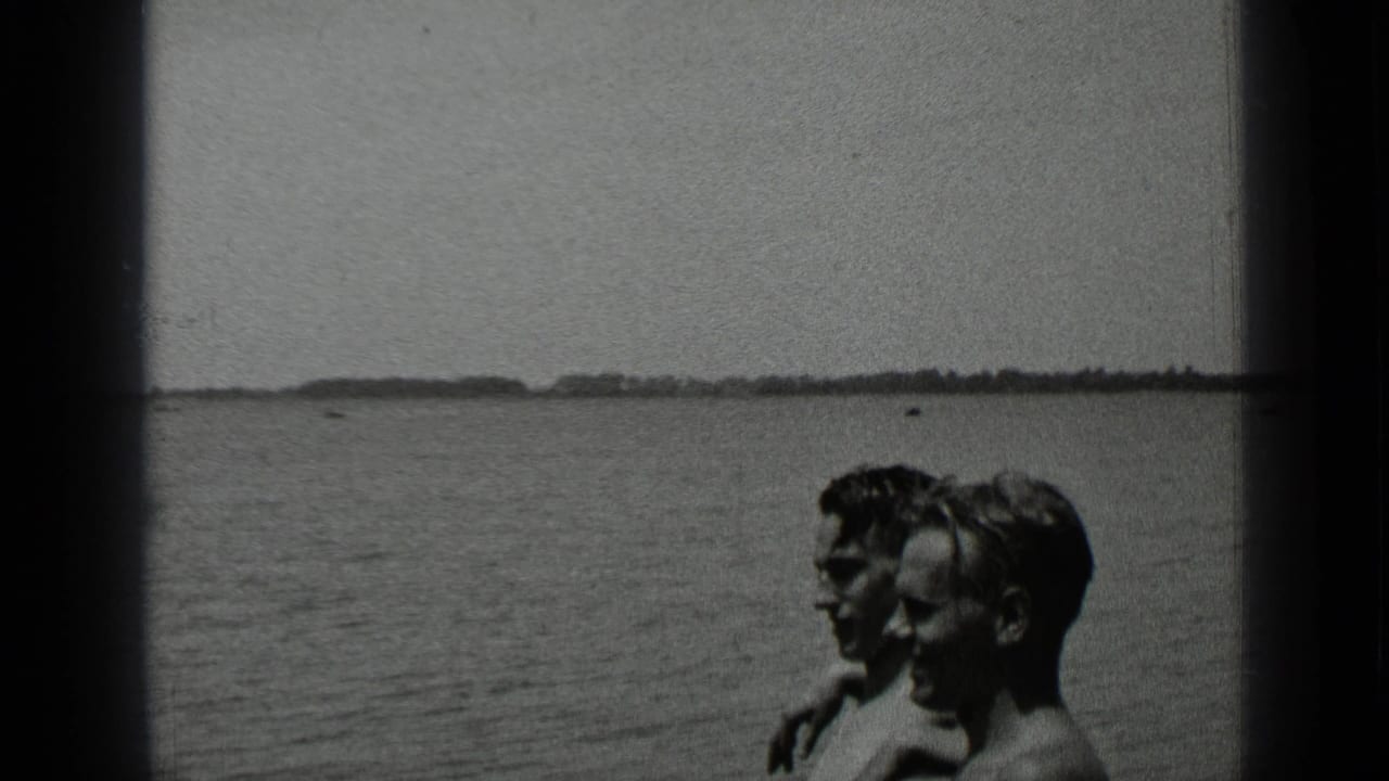 Two adults sit by a lake with open water and a distant shoreline behind them.