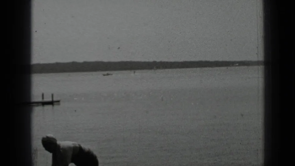 Black-and-white Clear Lake frame showing calm water, a small dock, distant shoreline, and a partially visible person near the lower edge.