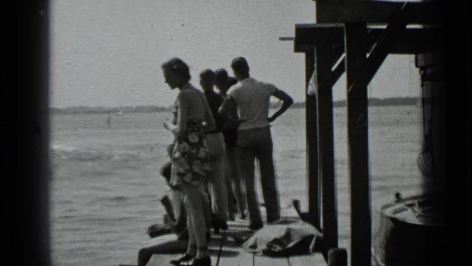 Clear Lake home-movie frame showing adults standing and sitting on a wooden dock while facing the water and distant shore.