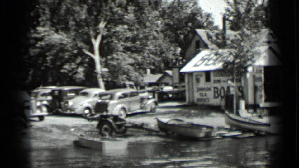 Clear Lake, California home-movie frame from 1939 showing a boathouse with a BOATS sign, parked late-1930s cars, boats, trees, and shoreline water.