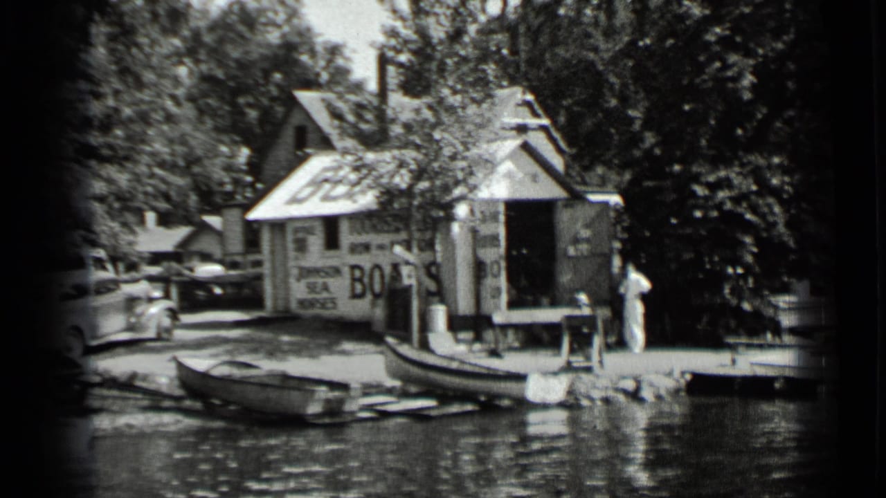 1939 Clear Lake frame showing the boat-rental area with a painted BOATS sign, rowboats along the shore, a parked car, dock edge, and a person nearby.