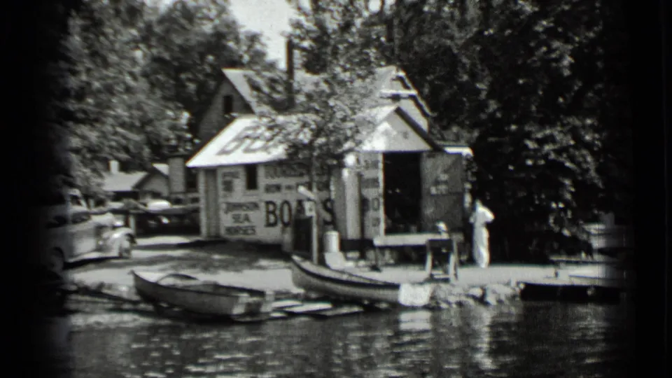 1939 Clear Lake frame showing the boat-rental area with a painted BOATS sign, rowboats along the shore, a parked car, dock edge, and a person nearby.
