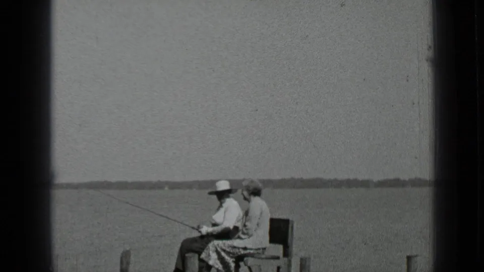 Clear Lake, California home-movie frame from 1939 showing two adults seated on a shoreline bench or dock, one holding a fishing rod over the lake.