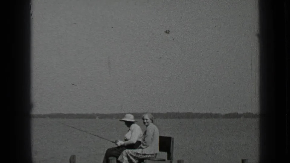 1939 Clear Lake frame showing two adults seated at the shoreline, with a fishing rod extending over calm water and a low distant shore in the background.