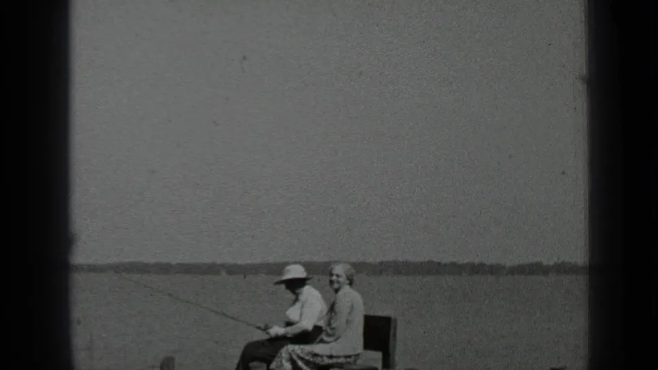 1939 Clear Lake home-movie frame of a couple on a lakeside bench, the man in a wide-brimmed hat holding a fishing rod.