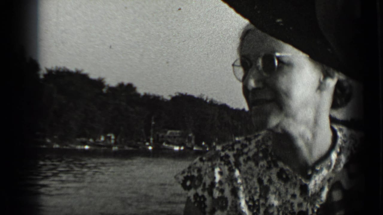 Elderly woman in glasses seated on a boat with lake and wooded shoreline behind her.