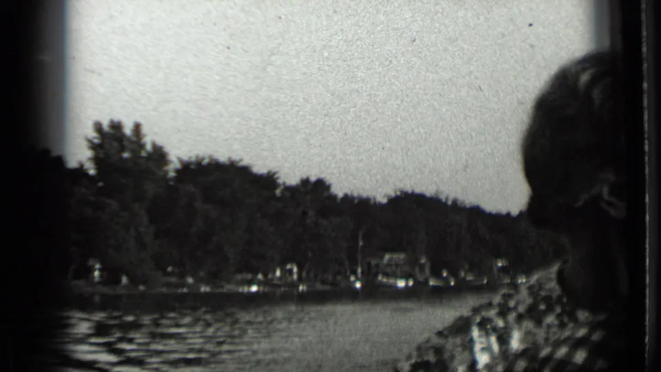 Home-movie frame from Clear Lake with a partially silhouetted passenger at right and small shoreline structures across the water.