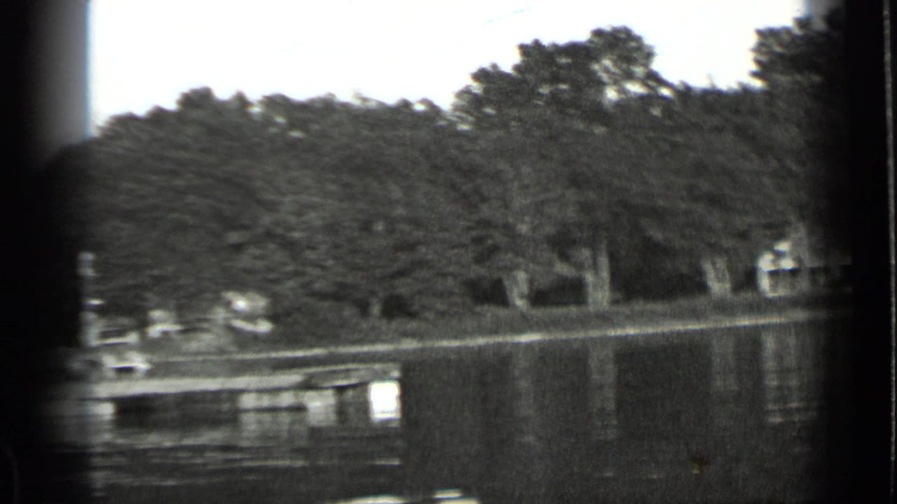 1939 Clear Lake view with calm water, dense trees, a low dock, and small buildings along the opposite shore.
