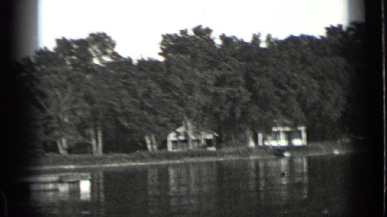 Black-and-white 8mm frame of Clear Lake shoreline, showing trees, modest lakeside buildings, and still water reflections.