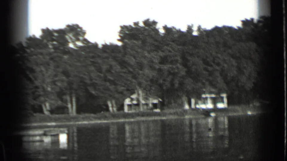 Black-and-white 8mm frame of Clear Lake shoreline, showing trees, modest lakeside buildings, and still water reflections.