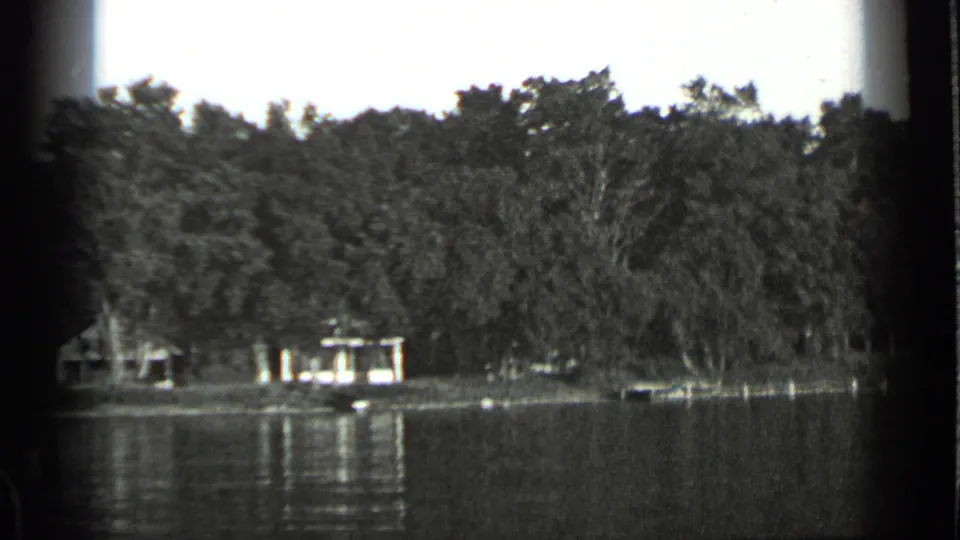 1939 Clear Lake home-movie frame with wooded banks, low shoreline structures, still water, and a small boat near shore.