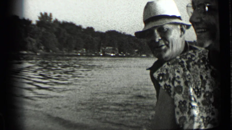 Man in a light fedora and patterned garment near Clear Lake, with another smiling adult partly visible at the frame edge.