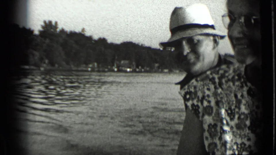 Man in a light hat smiles beside a partly cropped woman, with Clear Lake water and trees behind them.