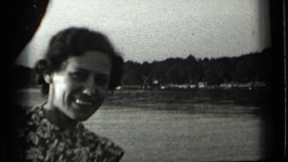 A smiling woman in patterned clothing faces the camera beside Clear Lake, California, with water and a wooded shoreline behind her.