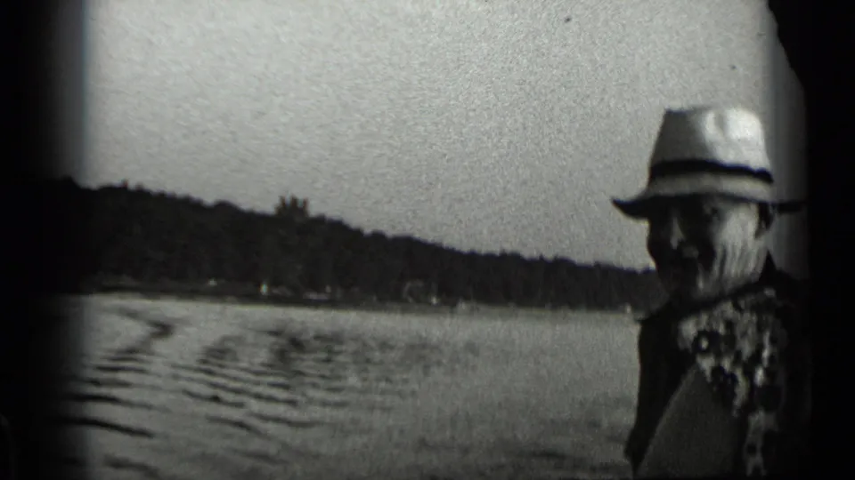 A man wearing a light-colored fedora-style hat and patterned shirt stands near Clear Lake with rippled water and shoreline behind him.