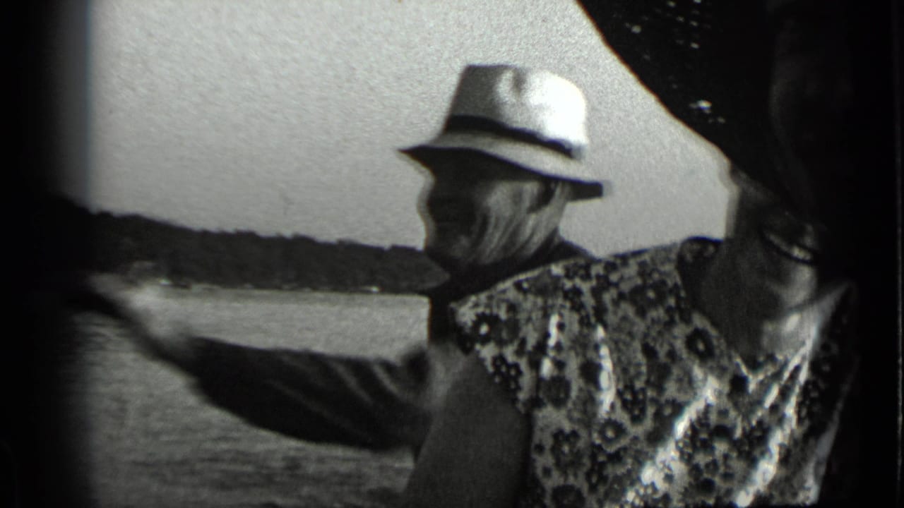 Older man in a light hat smokes beside a woman on a boat with lake water behind them.