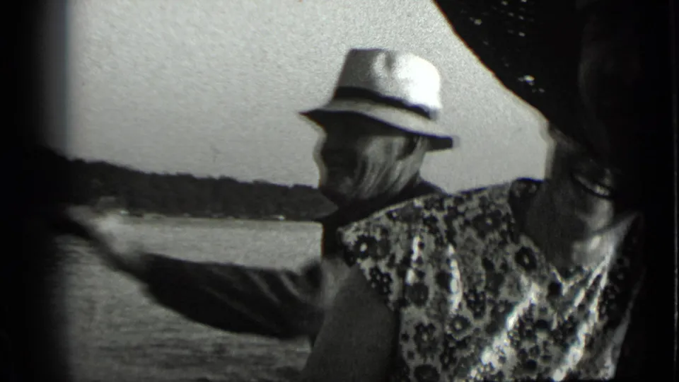1939 Clear Lake home-movie frame showing an older man in a light fedora smoking near a woman as shoreline and water pass behind them.