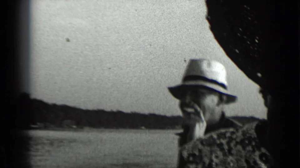 1939 Clear Lake frame of an older man in a light hat smoking on a moving boat, with water and wooded shoreline in the background.