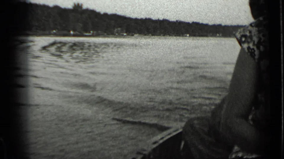 1939 Clear Lake frame showing wake trails on the water, wooded shoreline, and a partial passenger at the edge of the home-movie frame.