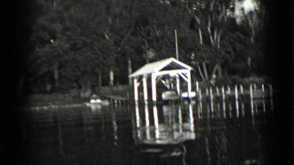 1939 Clear Lake home-movie frame showing a covered boathouse, dock pilings, shoreline trees, and water reflections.
