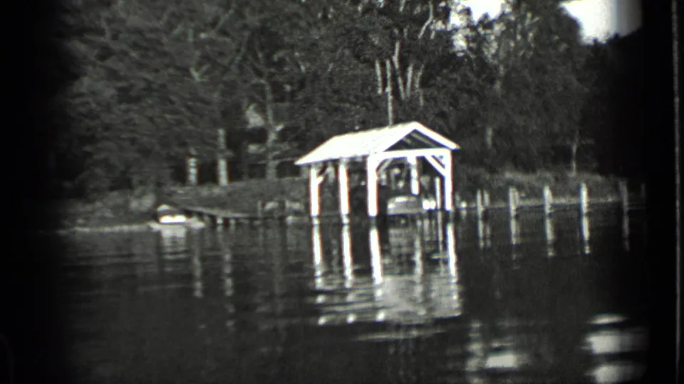 Black-and-white 8mm frame of a covered lakeside dock at Clear Lake, with pilings, trees, and rippled reflections.