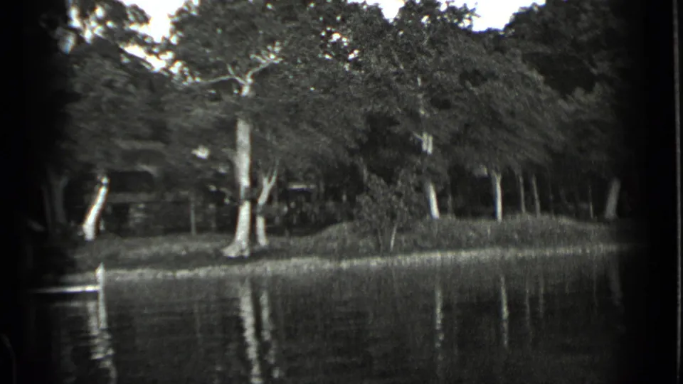 Black-and-white 8mm frame of the Clear Lake bank, with high water below trees and shoreline structures obscured by dense foliage.