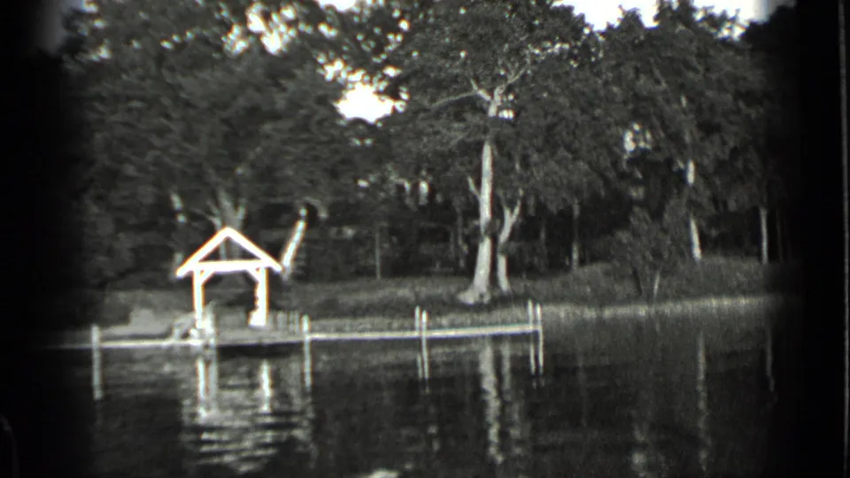 Black-and-white 8mm frame from Clear Lake showing the covered dock shifted farther right, with trees and still water around it.