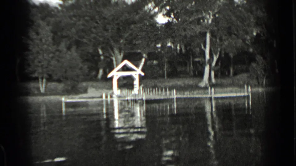 Vintage Clear Lake frame showing a small covered dock, pier posts, dense trees, and rippled reflections in black-and-white 8mm.