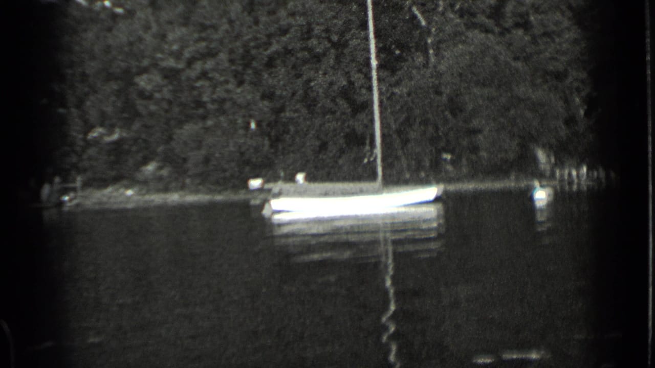 Small sailboat moored near a tree-lined lakeshore on calm water.