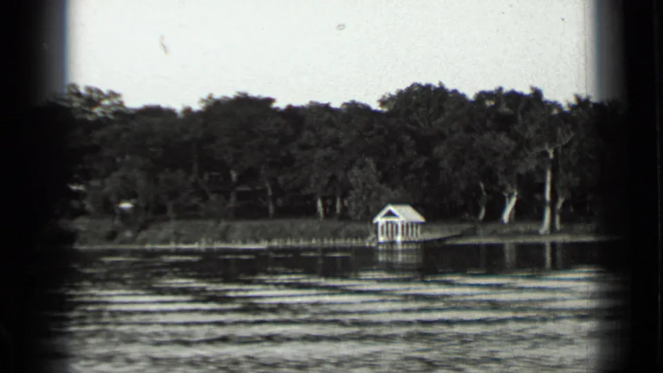 1939 Clear Lake view from the water, with a small white shore structure, reeds, trees, and horizontal ripples in the foreground.