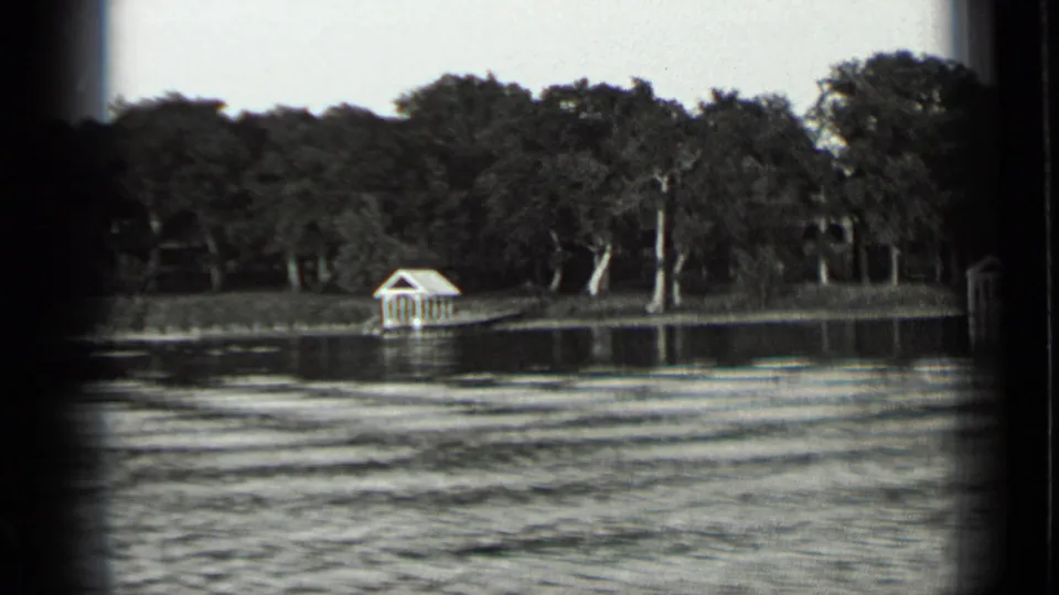 Clear Lake shoreline in black-and-white 8mm footage, showing trees, a small white boathouse or dock shelter, and textured water.