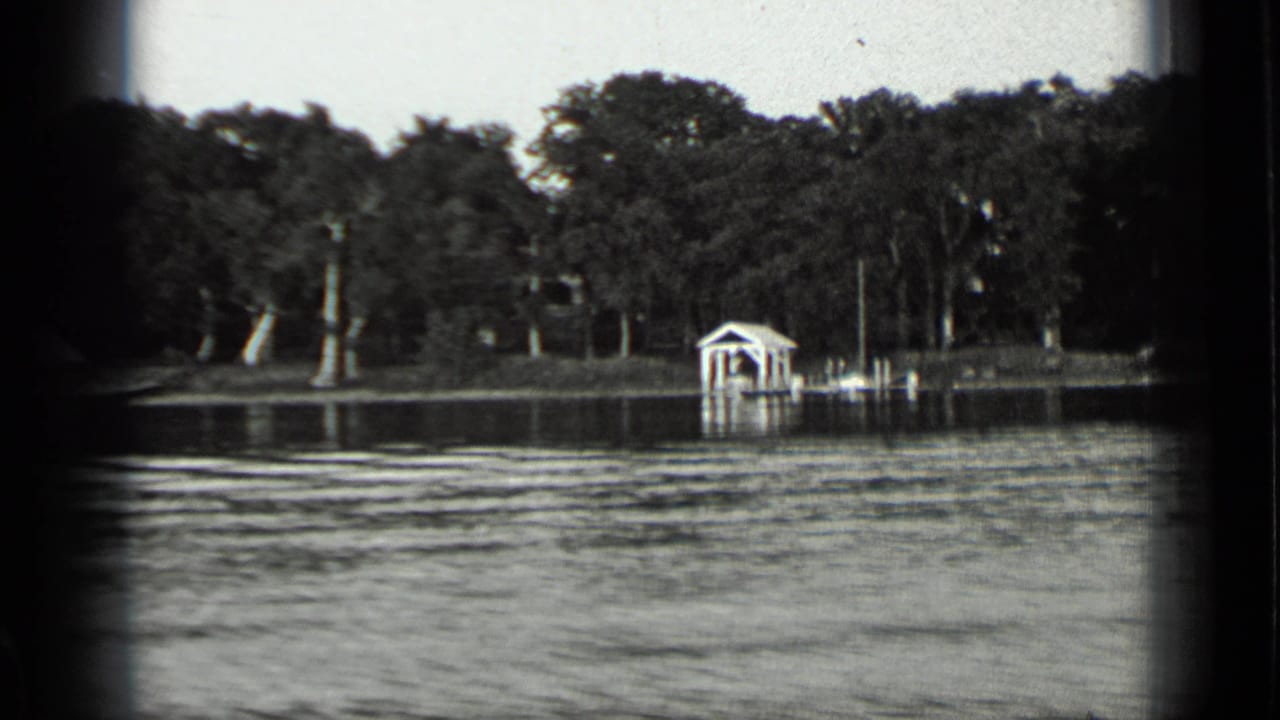 1939 Clear Lake shoreline view with rippled water, dense trees, a small boathouse, and a dock seen from a moving boat.