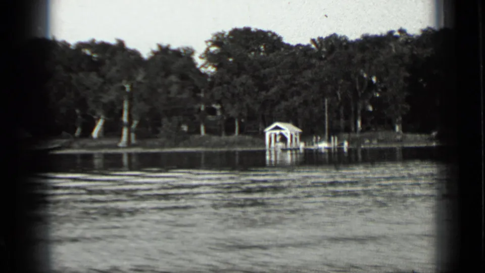 1939 Clear Lake shoreline view with rippled water, dense trees, a small boathouse, and a dock seen from a moving boat.