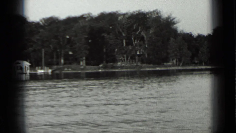 1939 Clear Lake boat-view frame showing rippled water, dense trees, a dock, and small shore structures partly obscured by the film edge.