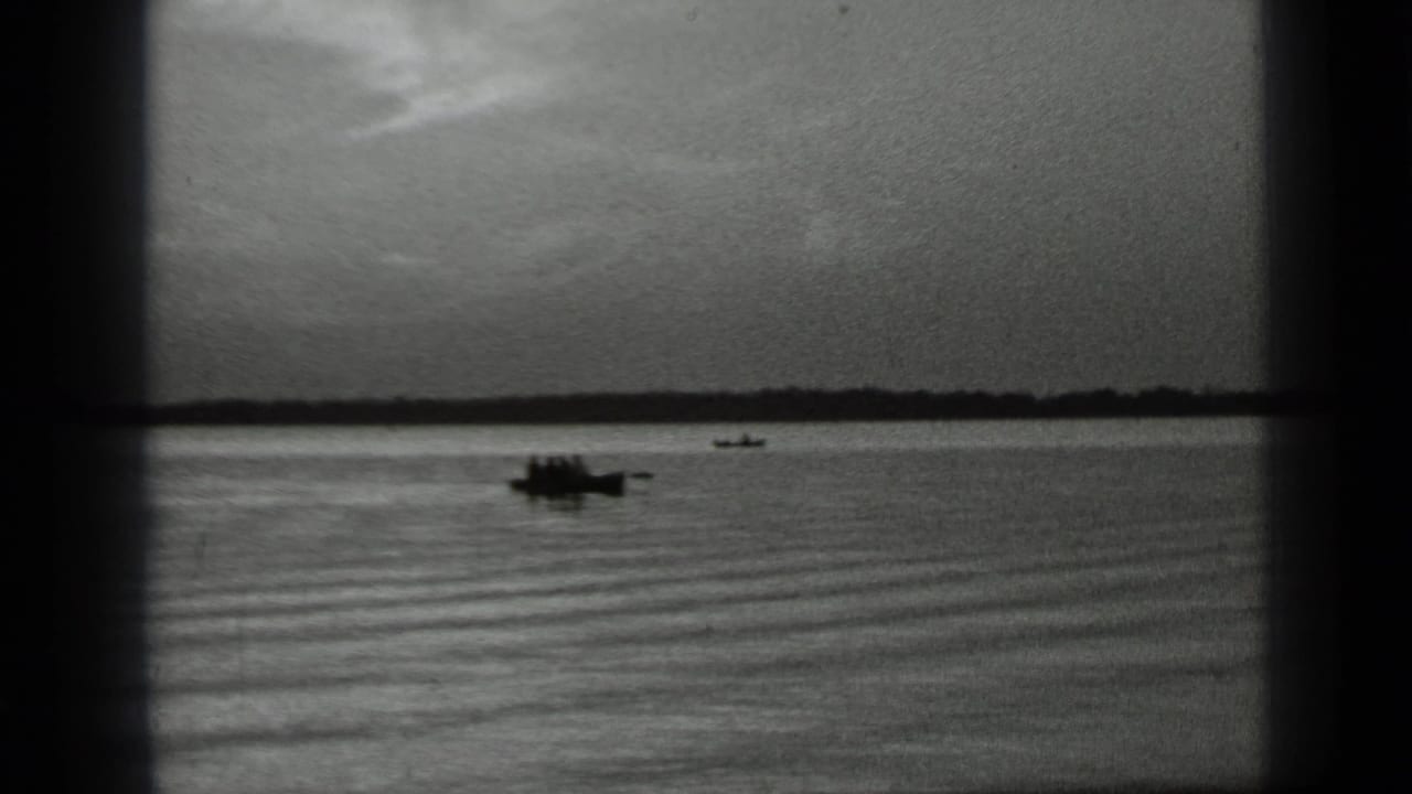 Distant small boat on calm lake water with a low shoreline under a cloudy sky.