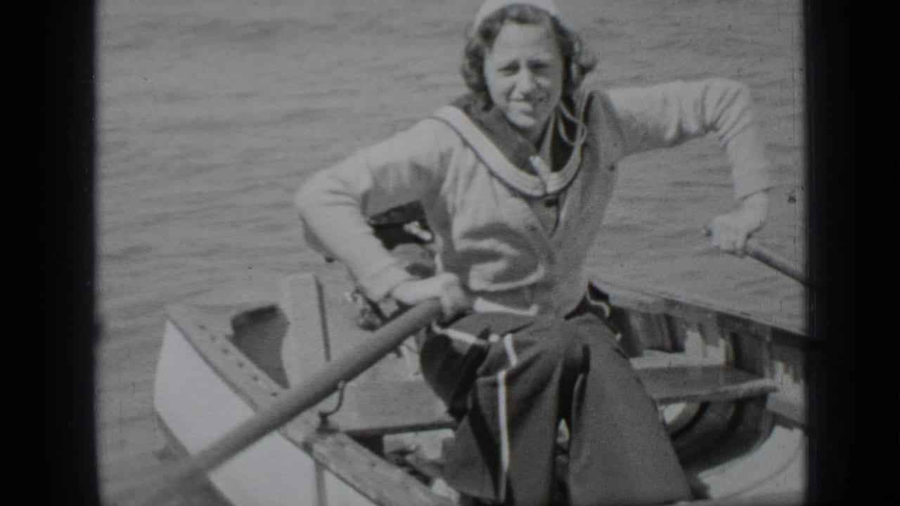 Woman in a sailor-style outfit seated in a small rowboat, holding an oar and facing the camera.