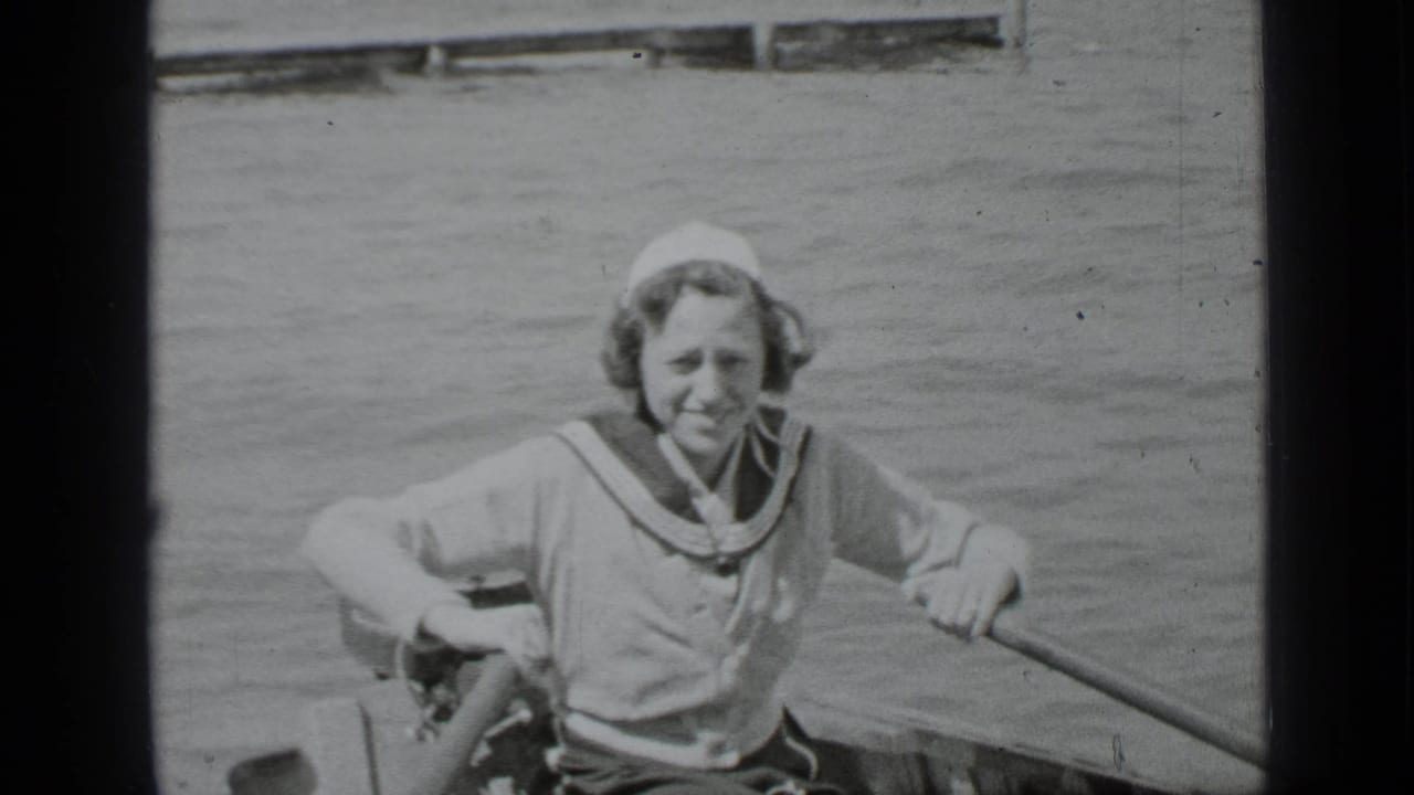 Young person in sailor-style clothing rows a canoe on lake water.
