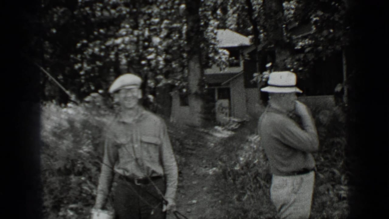 Two men stand in a wooded area near a cabin, one holding fishing gear.