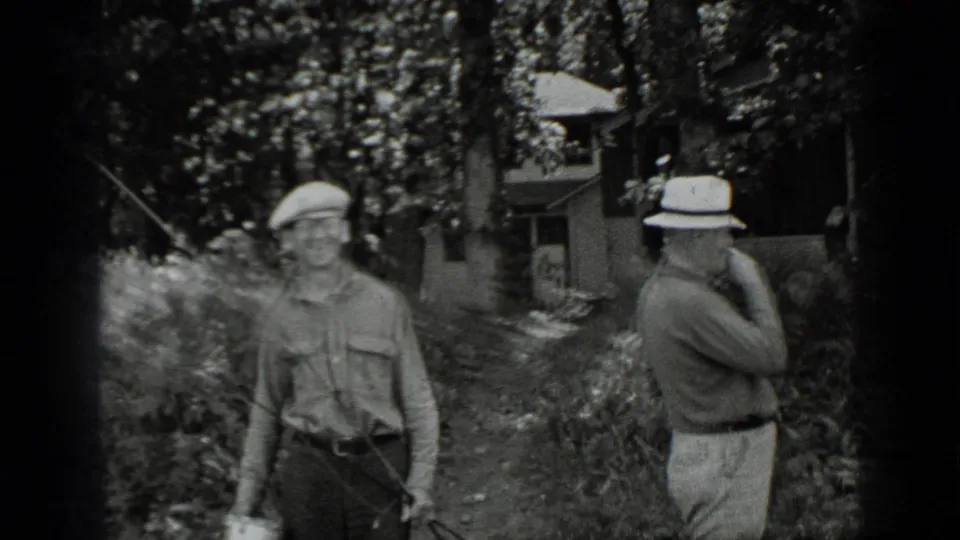Two men in hats stand along a wooded path near a lakeside cabin at Clear Lake, one with fishing gear and one smoking.